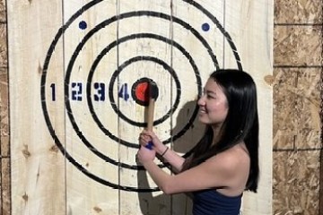 Person in front of a wooden target holding an axe with a bullseye hit.